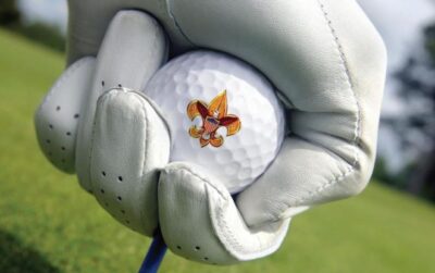A gloved hand pushes a golf ball and tee into the turf. The ball has the Scouting fleur-de-lis logo printed on it.