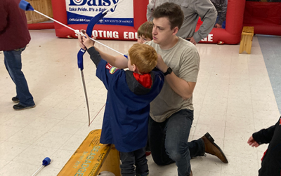 Tiny Cub Scout aiming a bow and arrow toward a target, assisted by a kneeling adult