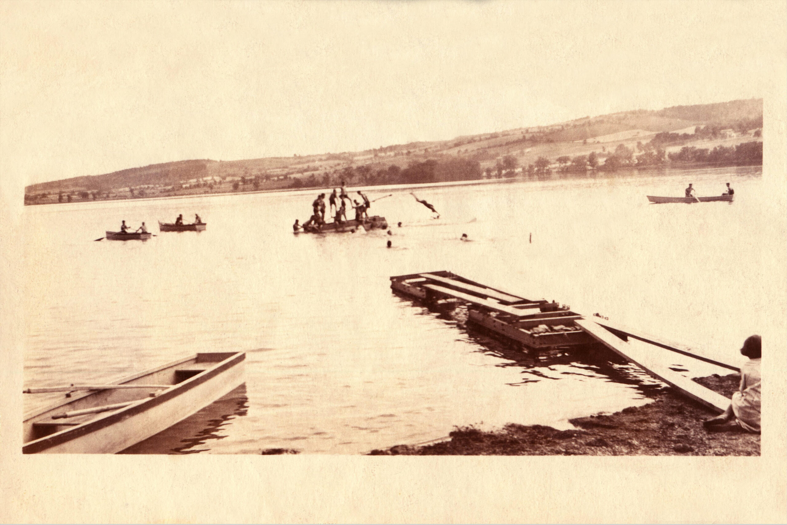 Monochrome image of Waneta Lake at Camp Gorton's waterfront showing campers on boats and a makeshift floating dock